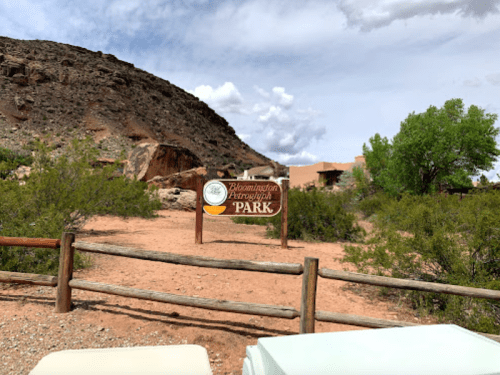 Sign for Bloomington Petroglyph Park, surrounded by rocky terrain and greenery under a cloudy sky.