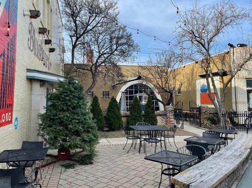 Outdoor patio with tables, chairs, and trees, featuring string lights and a mural on the wall in the background.