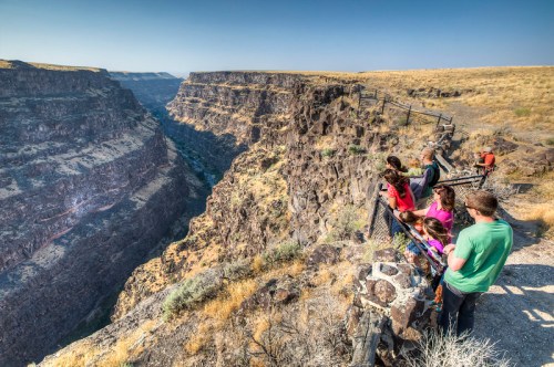 A group of people stands at a railing, overlooking a deep canyon with rocky cliffs and sparse vegetation.