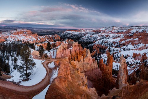 A panoramic view of Bryce Canyon, showcasing red rock formations and snow-covered trails under a colorful sky.