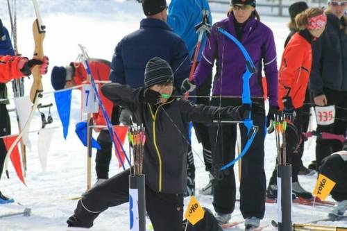 A person in winter gear aims a bow and arrow at a snowy archery range, surrounded by other competitors.