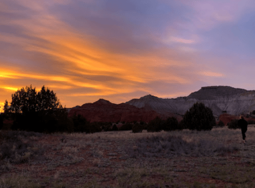 A serene landscape at sunset, featuring colorful skies, mountains, and a person walking in the distance.