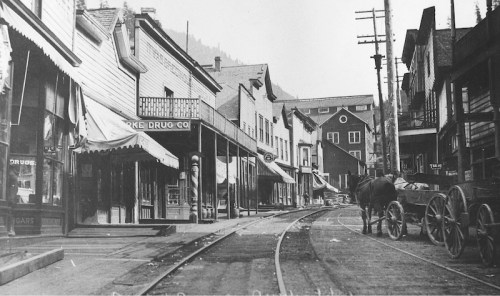 Historic street scene with wooden buildings, a horse-drawn wagon, and visible railway tracks in a small town.