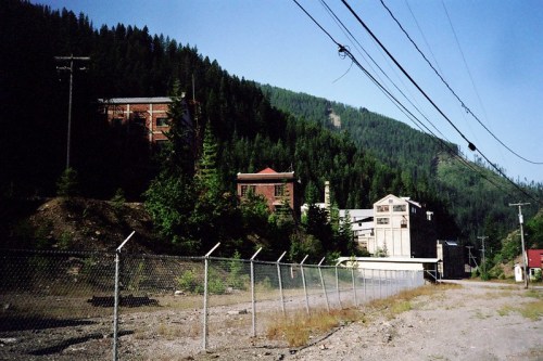 Abandoned buildings surrounded by trees and mountains, with a fence and power lines in the foreground.