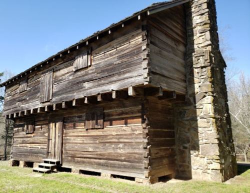 A rustic wooden cabin with a stone chimney, featuring multiple windows and a grassy area in front.