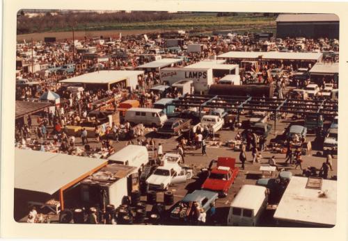 Aerial view of a bustling outdoor market filled with vendors, vehicles, and crowds of people browsing various goods.