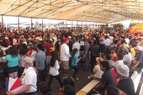A large crowd of people gathered under a tent, with colorful attractions in the background.