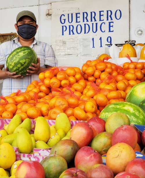 A vendor wearing a mask holds a watermelon in front of a colorful display of oranges, mangoes, and other fruits.