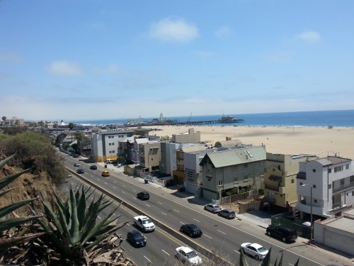 A coastal view featuring a beach, pier, and nearby buildings along a road under a clear blue sky.