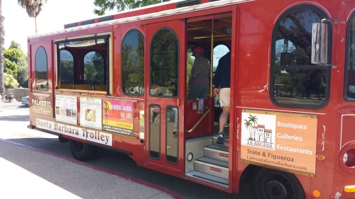 A red trolley labeled "Santa Barbara Trolley" parked with passengers boarding, featuring ads for local businesses.