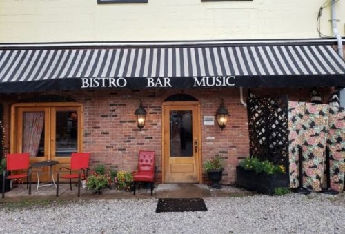 Exterior of a bistro with a striped awning, wooden door, and red chairs outside, featuring a brick facade and floral decor.