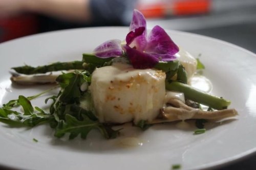 A gourmet dish featuring scallops, asparagus, and greens, garnished with a purple flower on a white plate.