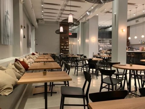 Modern restaurant interior with wooden tables, black chairs, and cozy cushions along the walls. Soft lighting creates a warm ambiance.