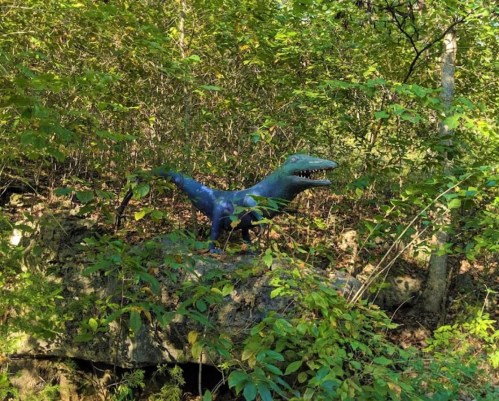 A colorful dinosaur statue partially hidden among lush green foliage in a forested area.