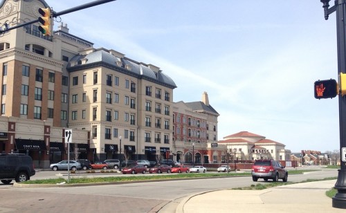 A street view of a modern urban area with shops, cars, and a traffic signal against a clear sky.