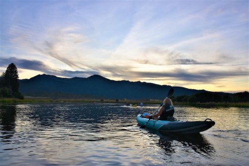 A person kayaking on a calm waterway with mountains and a colorful sky in the background during sunset.