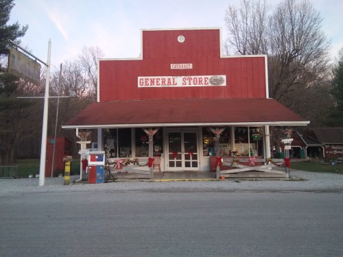 A red general store with a porch, decorated for the season, surrounded by trees and gravel road.