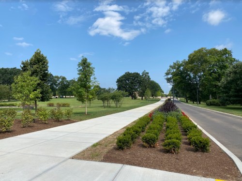 A paved path lined with greenery leads through a park under a clear blue sky.
