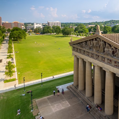 A wide view of a park featuring a replica of the Parthenon, with people enjoying the green space and city buildings in the background.