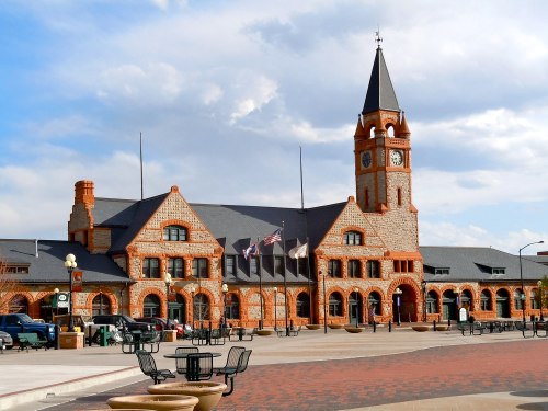 Historic brick building with a tall clock tower, flags flying, and a plaza in front. Blue sky with clouds above.