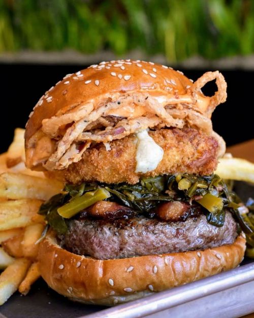 A close-up of a gourmet burger with a beef patty, fried onion rings, greens, and a sesame seed bun, served with fries.