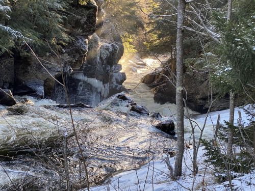 A serene winter scene of a river flowing through rocky terrain, surrounded by trees and soft sunlight filtering through.