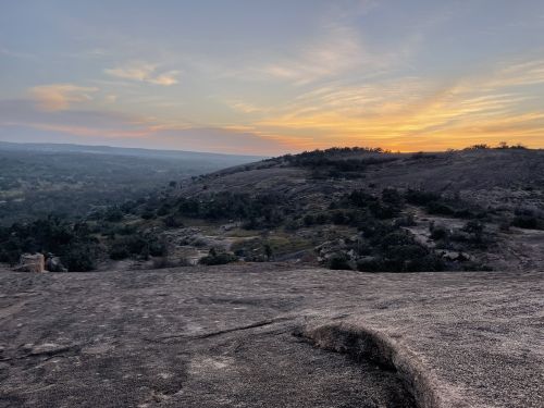 A scenic view of rolling hills at sunset, with a colorful sky and rocky terrain in the foreground.