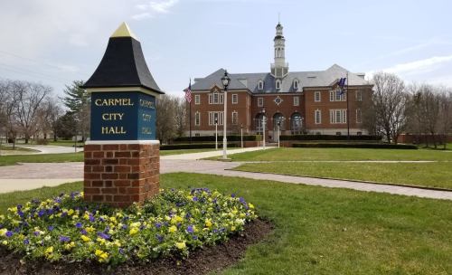 Carmel City Hall with a flower bed in the foreground, featuring a brick sign and a clear blue sky.
