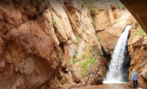 A waterfall cascades down rocky cliffs, with two people standing nearby, surrounded by lush greenery.