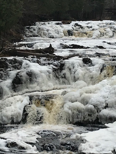 A cascading waterfall with frothy white foam and rocky terrain, surrounded by trees in a natural setting.