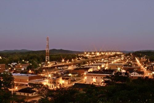 A panoramic view of a small town at dusk, with streetlights illuminating buildings and hills in the background.