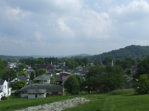 A scenic view of a small town surrounded by green hills and trees under a partly cloudy sky.