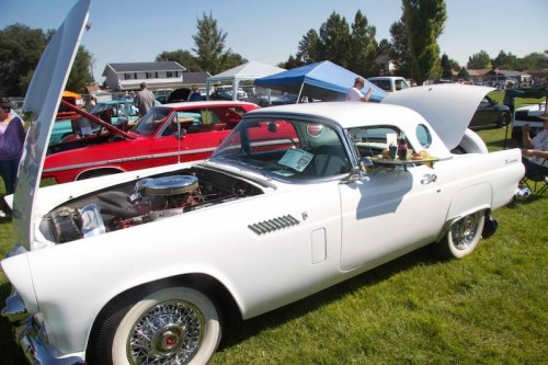 A classic white car with an open hood, showcasing its engine, parked on grass at a car show.
