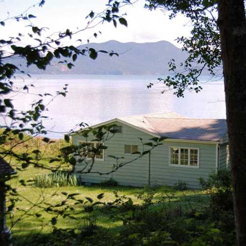 A serene lakeside view featuring a green house surrounded by trees and water in the background.