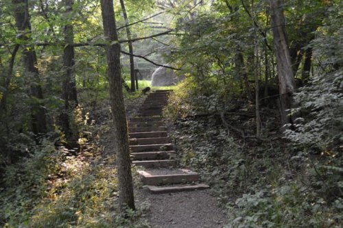 A stone staircase leads through a lush, green forest, surrounded by trees and dappled sunlight.