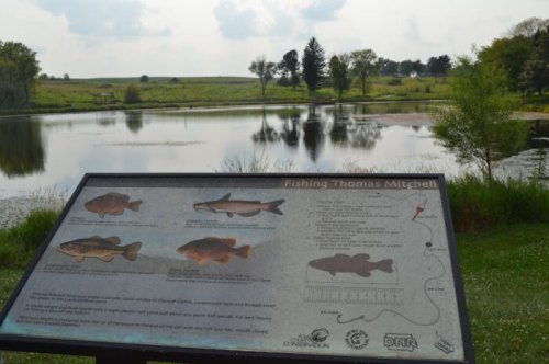 A sign about fishing at Thomas Mitchell Pond, with a scenic view of the water and surrounding greenery.