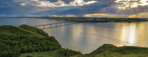 A scenic view of a river with a bridge, surrounded by lush green hills and dramatic clouds at sunset.