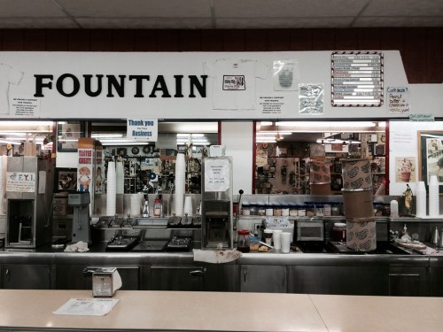 A retro diner counter with a "Fountain" sign, drink dispensers, and various memorabilia on the walls.