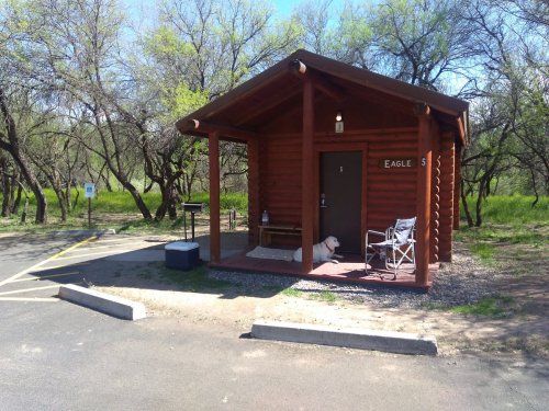 A small wooden cabin with a porch, surrounded by trees, featuring a bench and a chair outside.