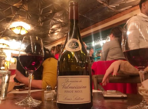 A bottle of Domaine de Valmoissine Pinot Noir 2016 on a table with two glasses of red wine and a lively restaurant background.