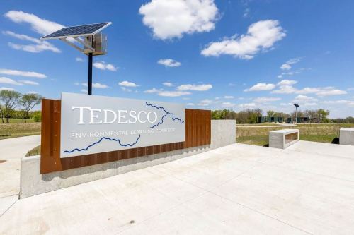 Sign for Tedesco Environmental Learning Corridor with solar panel, set against a blue sky and green landscape.