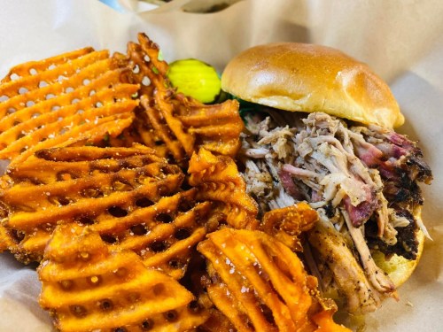 A plate featuring a pulled pork sandwich with a soft bun, accompanied by crispy waffle fries and a pickle.