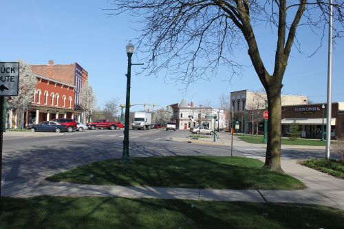 A small town intersection with trees, parked cars, and buildings under a clear blue sky.