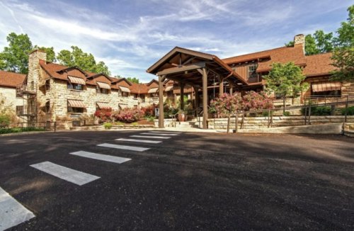 A rustic stone building with a wooden entrance, surrounded by greenery and a crosswalk in the foreground.
