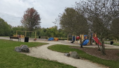 A playground with slides and climbing structures, surrounded by trees and grassy areas, under a cloudy sky.