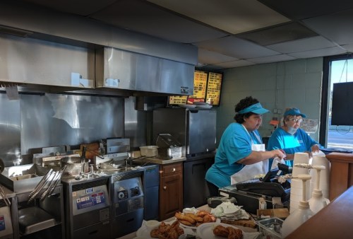 Two workers in blue shirts prepare food in a busy kitchen with cooking equipment and a menu display in the background.