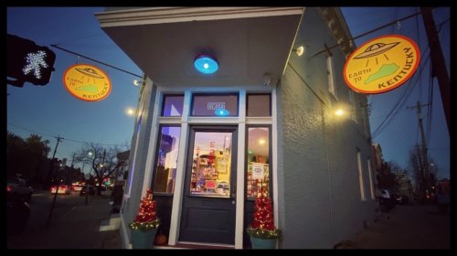 A storefront with colorful signs reading "Earth to Kentucky" and festive decorations, illuminated at dusk.