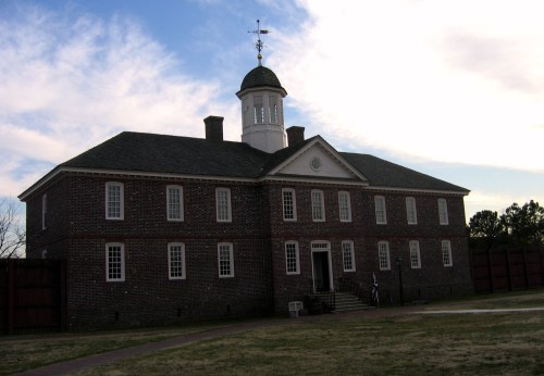 Historic brick building with a cupola, set against a cloudy sky, surrounded by grassy grounds.