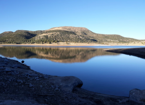 A serene lake reflects a mountain under a clear blue sky, surrounded by trees and rocky shoreline.