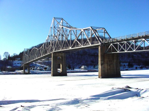 A metal bridge spans a frozen river, surrounded by snow-covered land and clear blue skies.